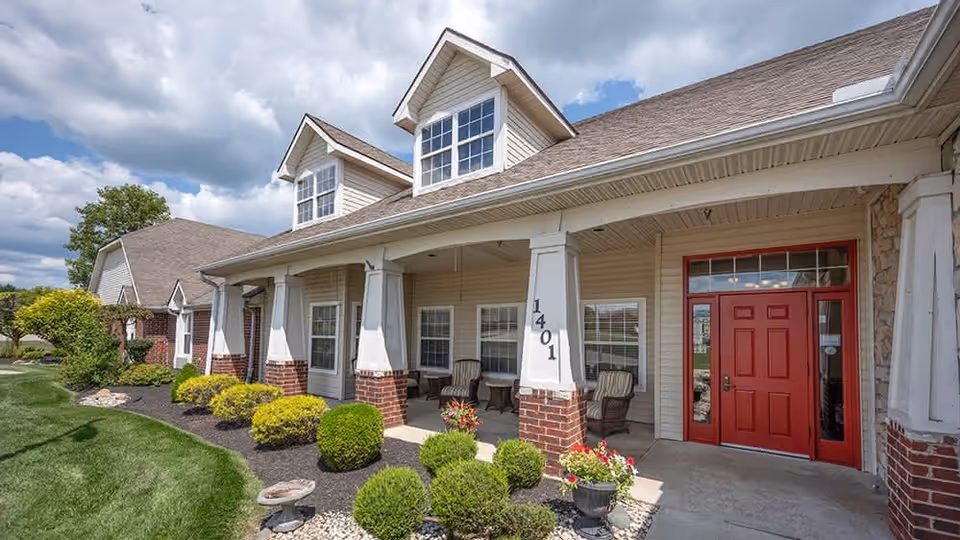 Exterior view of a single-story building with beige siding, red brick accents, and a red front door. The building has a covered porch with white columns and several chairs. The landscaping includes neatly trimmed bushes, a small flower bed, and a green lawn under a partly cloudy sky.