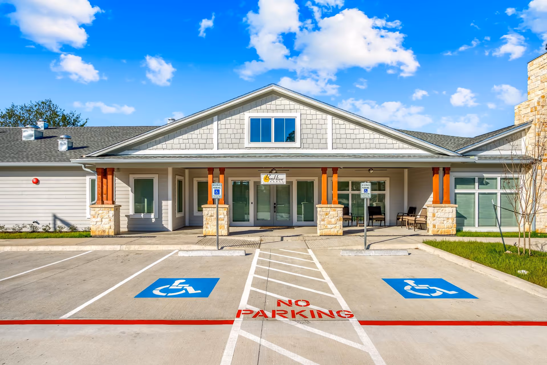 Front exterior view of BeeHive Homes of Spring building with a covered entrance, two handicap parking spaces, and a clear blue sky with some clouds.