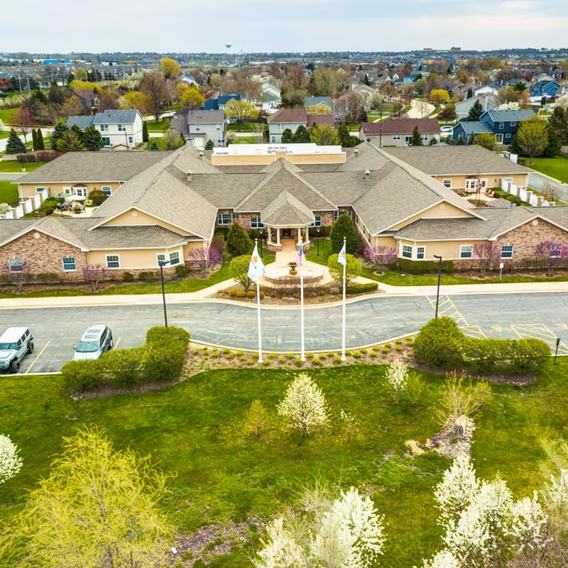 Aerial view of a senior living facility's exterior with a large beige building, flagpoles, and landscaped front lawn.