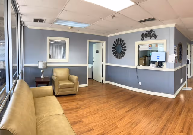 A waiting area with a beige leather sofa and armchair, a small wooden side table with a lamp, light blue walls with white trim, a window with a counter and computer monitor, a decorative round mirror, and a sign that says 'gather'. The floor is wooden and there is a hallway with open doors in the background.