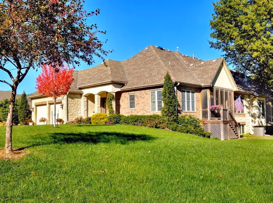 A single-story residential building with a combination of brick and stone exterior walls, a brown shingled roof, and a well-maintained green lawn in front. There are several trees, including one with red leaves and others with green foliage. The house features a screened porch with hanging flower baskets and an American flag displayed near the entrance. The sky is clear and blue.