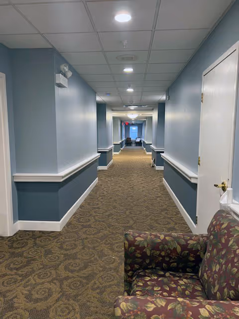 A well-lit hallway featuring blue walls, carpeted flooring, and a patterned armchair on the side.