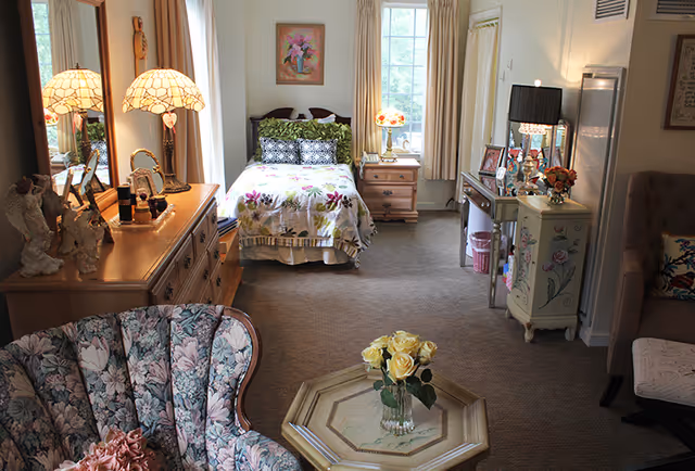 A cozy assisted living room with a single bed covered in floral bedding, a wooden dresser with two decorative lamps, a floral upholstered armchair, a small wooden coffee table with a vase of yellow roses, and a side table with a lamp and framed photos. The room has beige carpet and light-colored curtains on the windows.