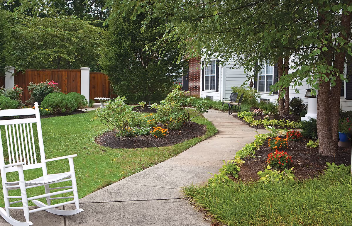 A peaceful outdoor garden area at Blue Bell Place featuring a concrete pathway winding through well-maintained flower beds with various shrubs and colorful flowers. There is a white rocking chair on the left side of the path and a black metal bench near the building entrance. The building exterior is light gray with white trim and large windows, surrounded by green trees and bushes.