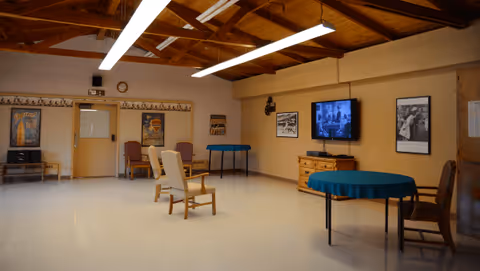 A spacious common room with wooden ceiling beams and fluorescent lighting. The room contains several chairs, two round tables covered with blue tablecloths, a wooden dresser with a flat-screen TV mounted above it, and framed black-and-white photographs on the walls. The floor is light-colored, and there is a door and a clock on the far wall.