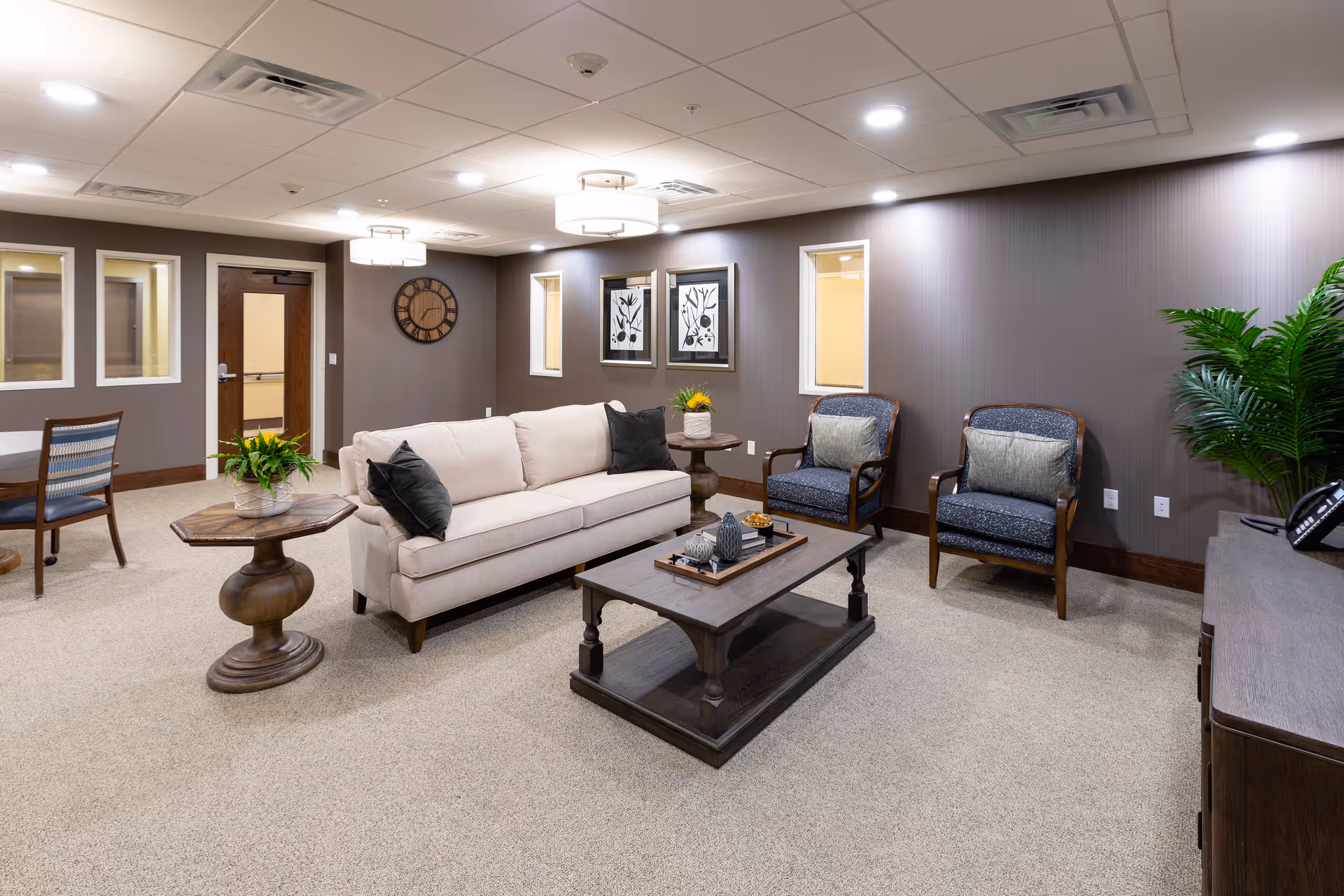 A cozy senior living common area with a beige sofa adorned with black pillows, two blue armchairs with gray cushions, a wooden coffee table with decorative items, and a round wooden side table with a green plant. The room has beige carpet, gray walls with framed artwork, a large wall clock, ceiling lights, and a potted plant near a wooden cabinet with a telephone.