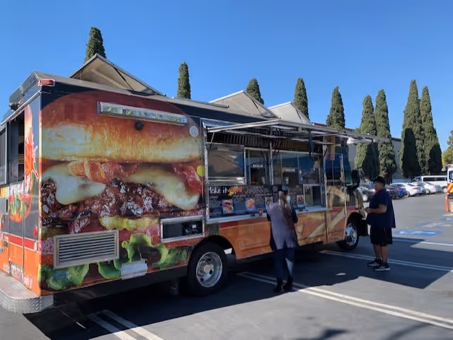 A colorful food truck parked in a sunny parking lot with two people at the service window and tall cypress trees in the background.