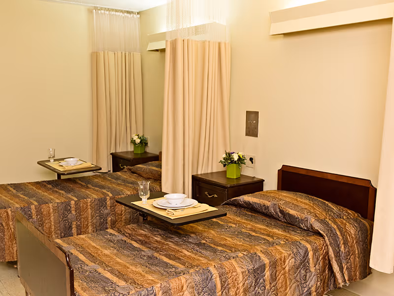 A room with two single beds separated by a curtain. Each bed has a brown patterned bedspread and a bedside table with a small green plant. There are trays with plates, bowls, napkins, and glasses placed on small tables over each bed.