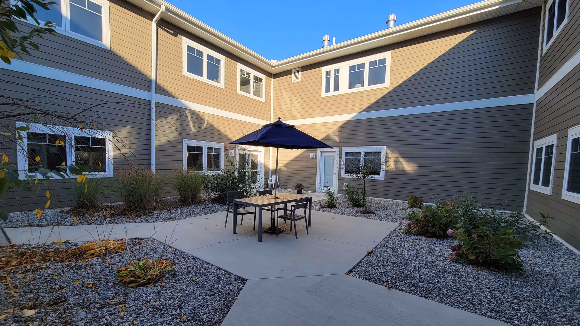 Outdoor courtyard area at Serene Gardens of Sterling Heights with a concrete patio featuring a table, four chairs, and a large blue umbrella. The courtyard is surrounded by a two-story building with multiple windows and landscaped with gravel and various plants.