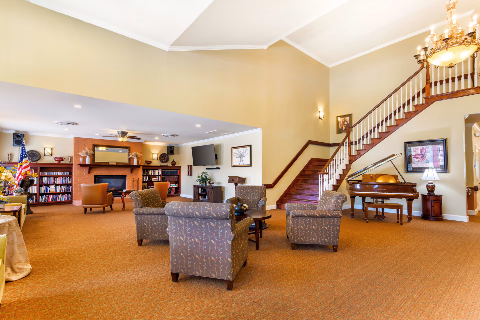 A spacious senior living facility common area with beige walls and carpet. The room features a seating arrangement with four patterned armchairs around a small round table. In the background, there is a brick fireplace flanked by bookshelves filled with books and decorative items. An American flag stands near the fireplace. To the right, a wooden staircase with white spindles leads to an upper floor, and a grand piano with a bench is positioned near the staircase. A chandelier hangs from the ceiling, and framed artwork decorates the walls.