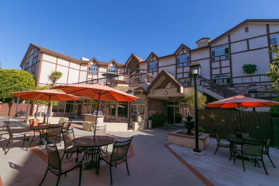 Outdoor courtyard with round tables, chairs and red umbrellas in front of a multi-story Tudor-style assisted living building under a clear blue sky.