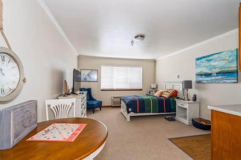 A cozy bedroom with a bed covered in a colorful striped blanket, a nightstand with a lamp, a blue armchair, a dresser with a TV, and a round table with a Scrabble game set up. The room has beige walls, a window with blinds, and a painting of a seascape on the wall.