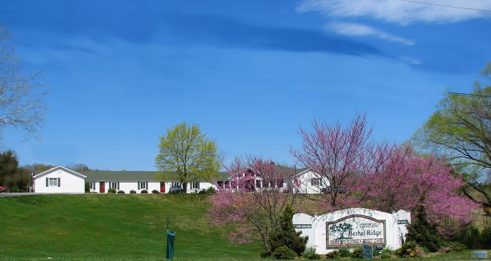 A single-story white building with a green roof situated on a grassy hill under a clear blue sky. In front of the building, there are trees with green and pink blossoms. A white sign in the foreground reads 'Bethel Ridge Assisted Living & Adult Care.'