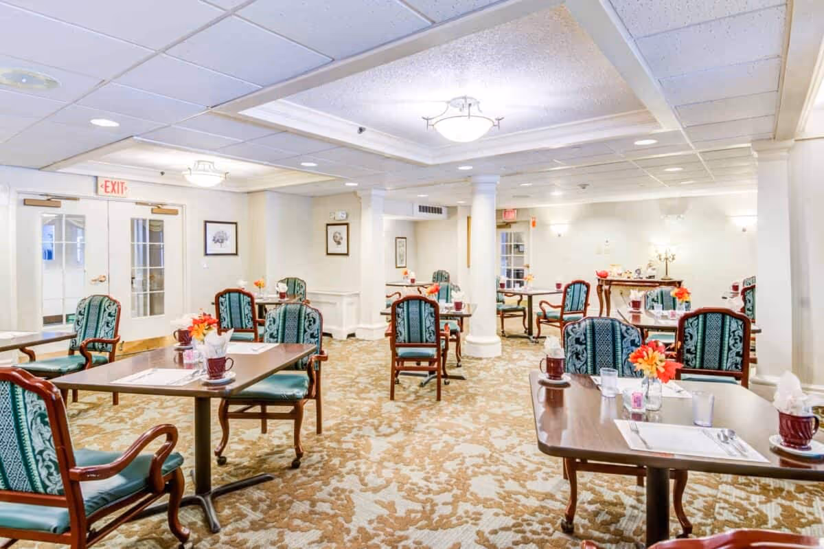 A bright and spacious dining room with multiple wooden tables and green upholstered chairs arranged neatly. Each table is set with placemats, napkins, cups, and small flower arrangements. The room features white walls, decorative columns, and a patterned carpet. There are framed pictures on the walls and exit doors with glass panels at the back.