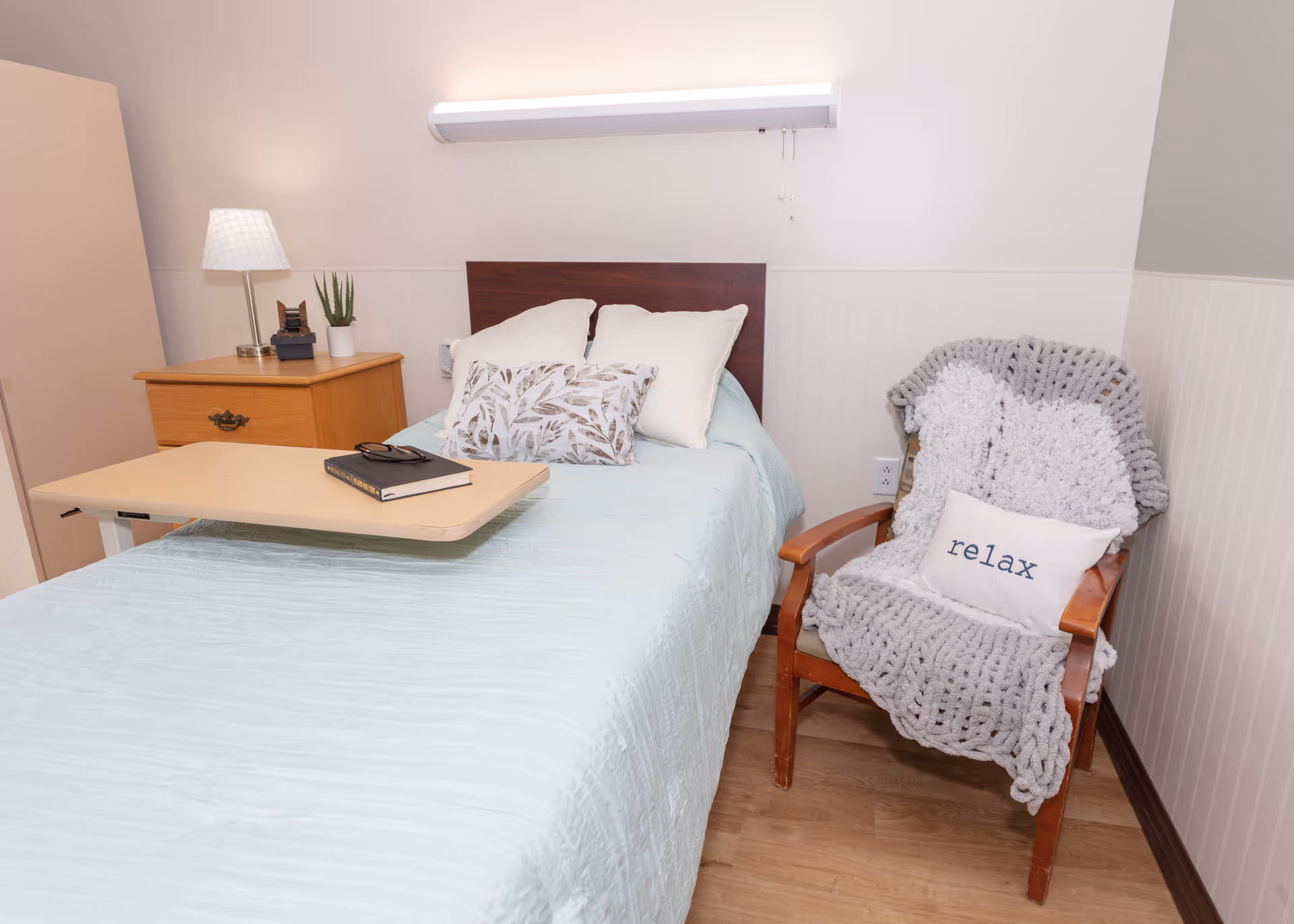 A cozy bedroom in a senior living facility featuring a single bed with light blue bedding and multiple pillows, a wooden bedside table with a lamp and small plant, and a wooden chair draped with a chunky gray knit blanket and a white pillow with the word 'relax' on it. The room has light-colored walls and wood flooring.