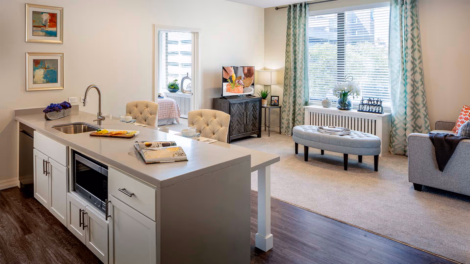 Open-plan apartment interior with a kitchen island and sink facing a furnished living room with a large window, ottoman, and sofa.