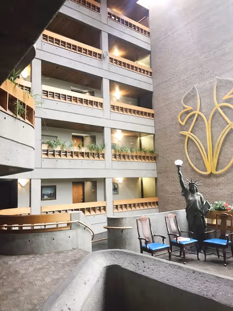 Interior multi-story atrium of an apartment building with open balconies, plants, seating, and a small Statue of Liberty sculpture.