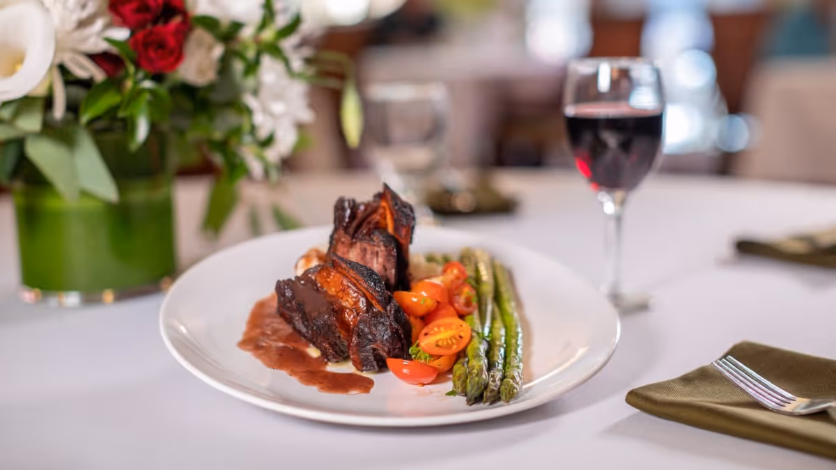 A plated meal featuring slices of cooked meat with sauce, cherry tomatoes, and asparagus on a white plate, set on a white tablecloth with a glass of red wine and a green napkin with a fork beside it. A floral arrangement with red and white flowers is visible in the background.