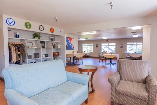 A spacious common area in a senior living facility with light blue and patterned armchairs, wooden side tables, and several round tables with chairs near large windows. The walls are decorated with military insignias and memorabilia, including jackets and helmets displayed on shelves.