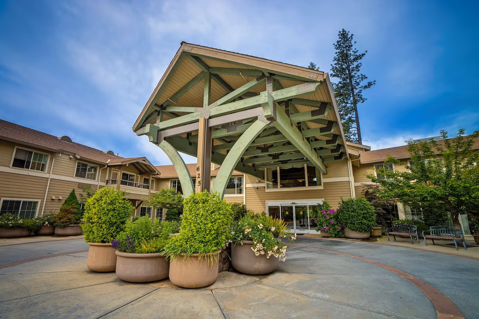 Entrance of Eskaton Village Grass Valley featuring a large covered porte-cochère with green wooden beams, surrounded by large potted plants and shrubs. The building has beige siding with white trim and multiple windows, under a partly cloudy blue sky.