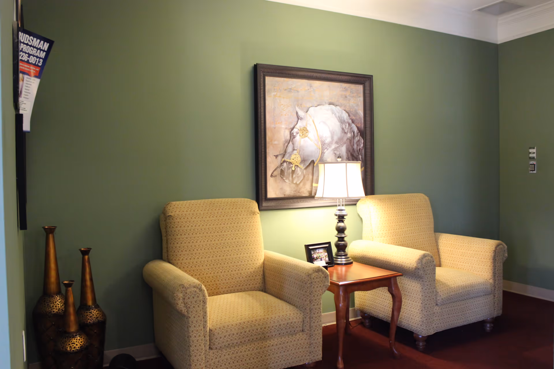 Two beige upholstered armchairs with a subtle pattern are placed on either side of a small wooden side table with a lamp and a framed photo. The wall behind is painted green and features a framed painting of a white horse's head. Three decorative tall vases are positioned on the floor to the left of the chairs.