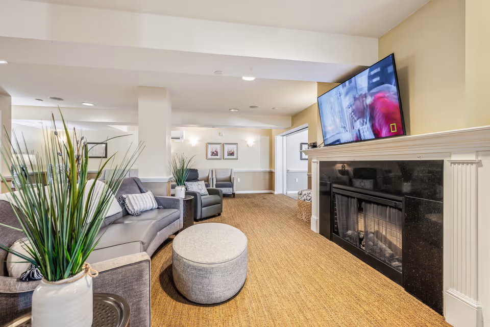 A cozy living room area in a memory care facility featuring a curved gray sofa with decorative pillows, a round gray ottoman, a black fireplace with a mounted flat-screen TV above it, and several armchairs. The room is decorated with tall green plants in white vases and framed artwork on the walls. The carpet is a warm brown color, and the walls are painted in neutral tones.