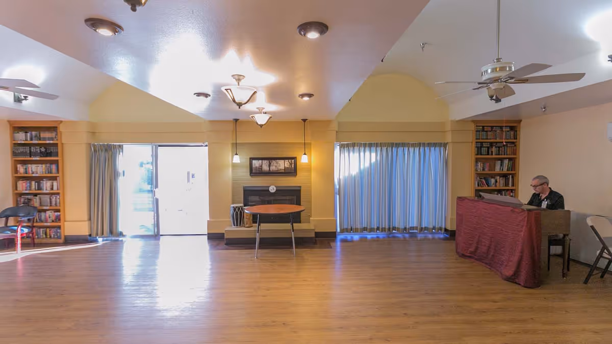 Spacious senior living common room with hardwood floor, fireplace, bookshelves, and a person seated at a piano.