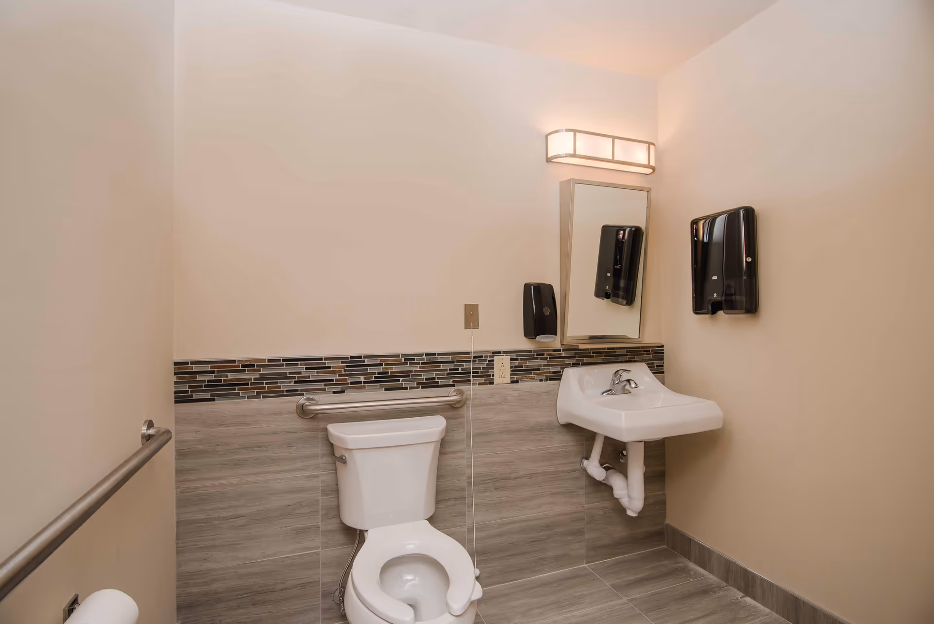 A clean and accessible bathroom with a white toilet and a wall-mounted white sink. The walls are beige with a decorative tile strip above the toilet and sink. There are metal grab bars on the left wall and behind the toilet. A rectangular mirror is mounted above the sink, with a light fixture above it. Two black soap or sanitizer dispensers are mounted on the wall near the sink.