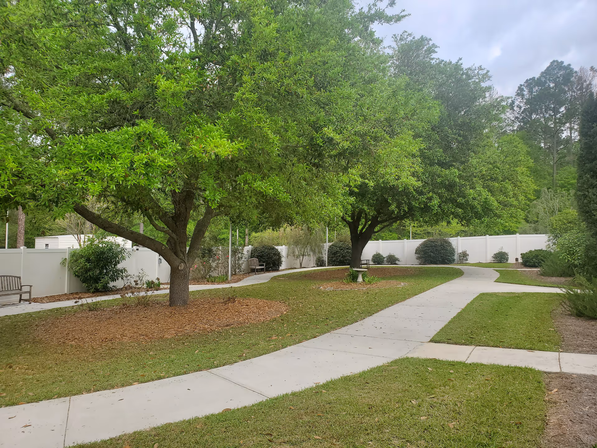 A peaceful outdoor garden area with a winding concrete pathway, green grass, and several large leafy trees. There are benches along the pathway and a white fence enclosing the space, with bushes and other greenery around the edges.