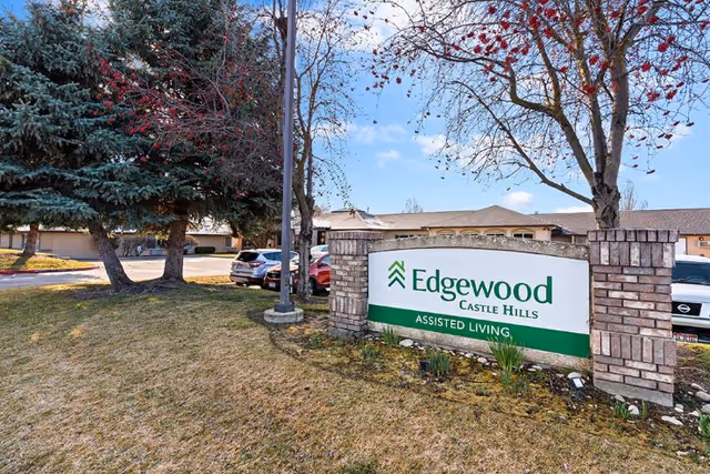 Outdoor view of the entrance sign for Edgewood Castle Hills Assisted Living facility, surrounded by grass, trees, and parked cars in the background under a partly cloudy sky.