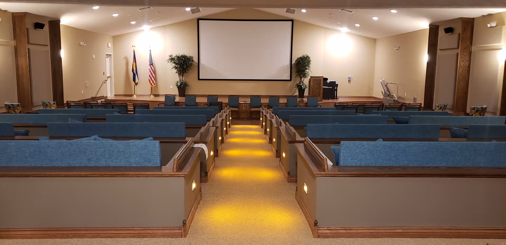 Interior view of a meeting or presentation room with rows of blue cushioned pew-style seating facing a stage with a large blank projection screen. Two flags and two potted plants are on the stage, with a podium and audio equipment on the right side.
