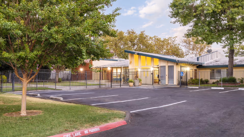 Gated entrance and empty parking lot in front of a single-story care facility with trees and a slanted roof.