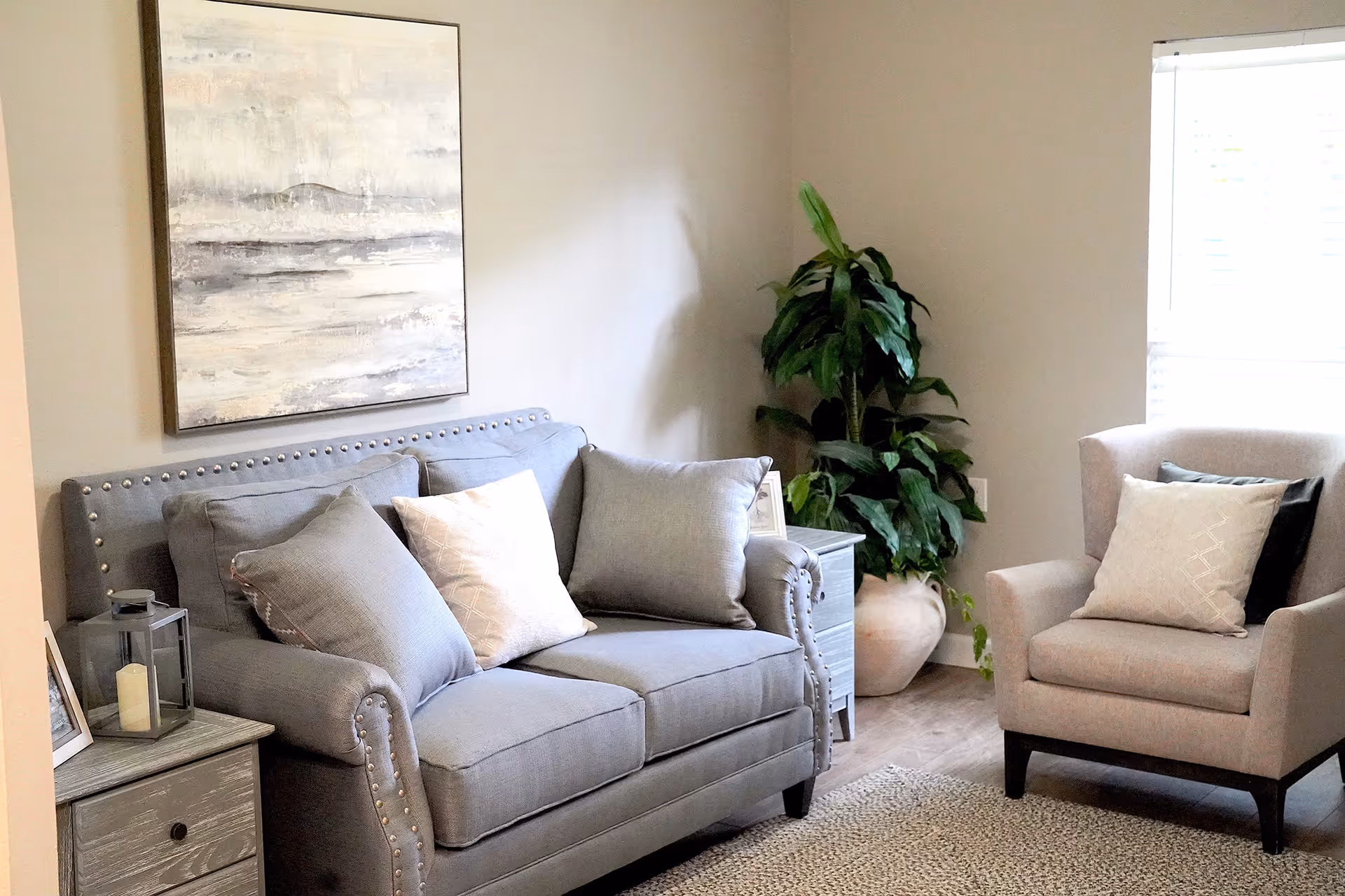 A cozy living room area with a gray upholstered loveseat adorned with three cushions, a beige armchair with a cushion, a large potted plant in the corner, a window with blinds, a side table with a candle lantern and framed photo, and a large abstract painting on the wall.