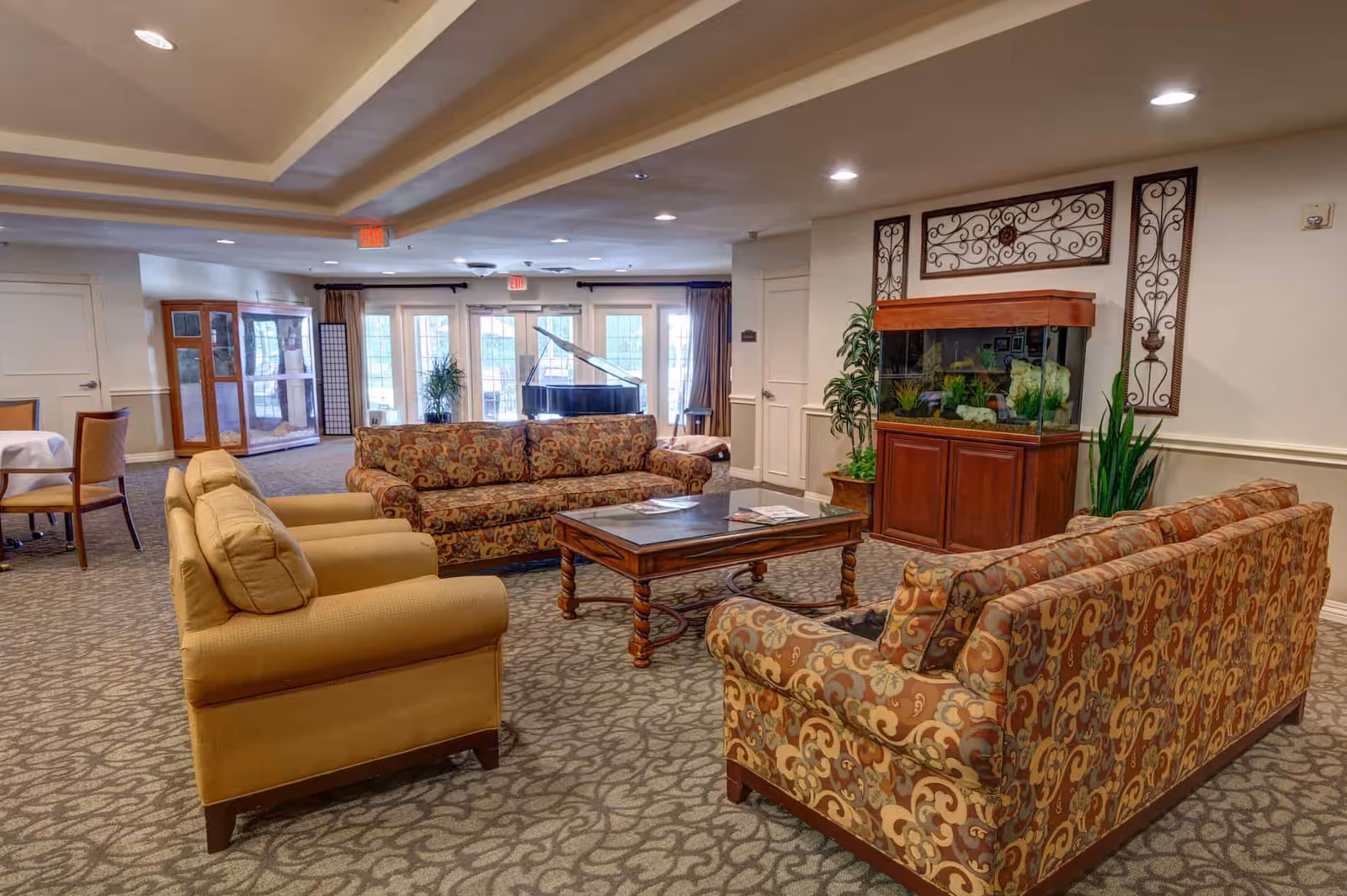 Spacious senior living common room with patterned sofas and armchairs around a coffee table, a large aquarium, and a grand piano by glass doors.