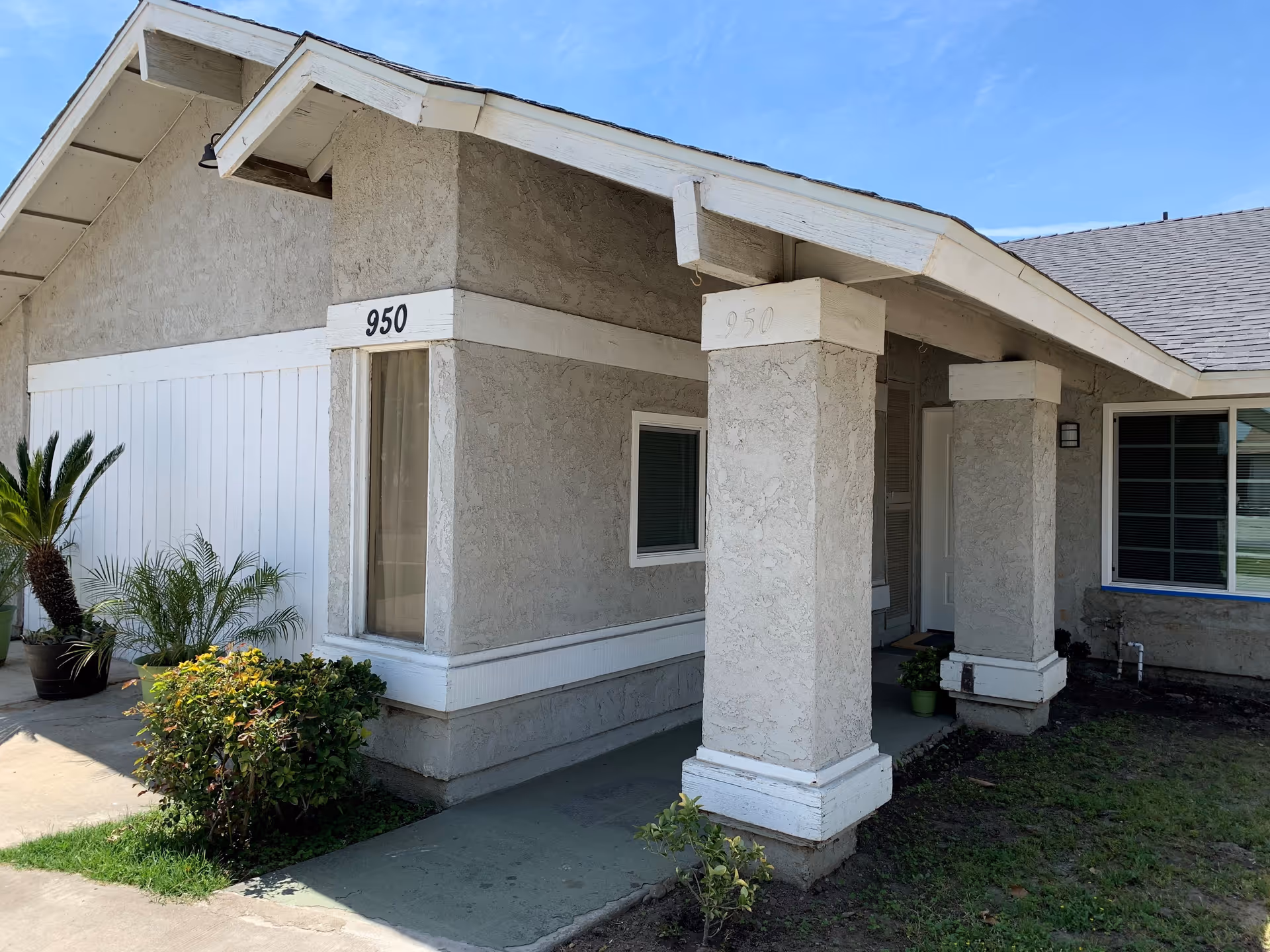 Exterior view of a single-story building with beige stucco walls and white trim. The building has two large square columns supporting a small porch roof. The number 950 is displayed on the corner of the building and on one of the columns. There are several potted plants and bushes near the entrance, and the sky is clear and blue.