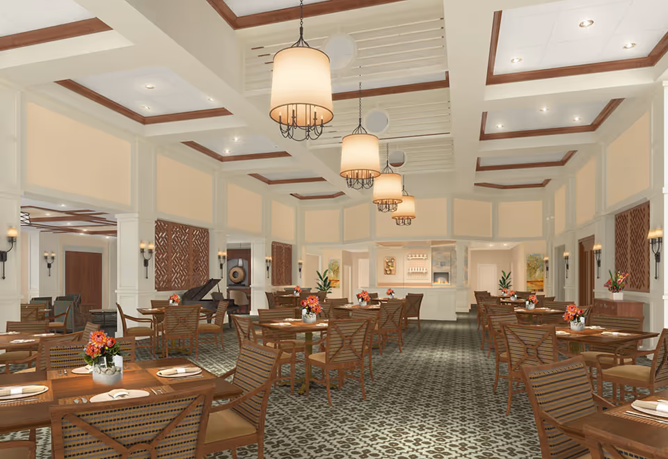 A spacious and elegant dining room with multiple wooden tables and chairs arranged neatly. Each table has a small flower arrangement and place settings with napkins. The room features high ceilings with recessed lighting and hanging pendant lamps. The walls are decorated with wooden panels and sconces, and there is a fireplace and artwork visible in the background.