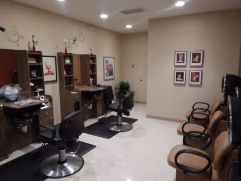 Interior view of a salon area with three styling stations, each equipped with a mirror, countertop, and salon chair. On the right side, there are three brown chairs lined up against the wall. The walls are beige with framed pictures and decorative wall art. The floor is light-colored tile.
