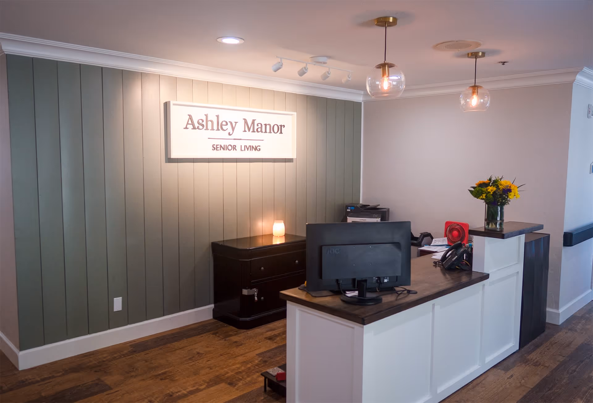 Reception area of Ashley Manor Senior Living with a wooden desk, computer monitor, telephone, and a vase of flowers. The wall behind the desk has vertical green paneling and a sign that reads 'Ashley Manor Senior Living'. Two pendant lights hang from the ceiling.