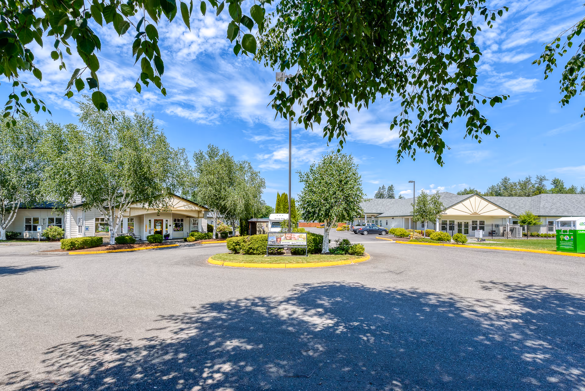 Exterior view of Heritage House Buckley Assisted Living & Memory Care facility on a sunny day with blue sky and scattered clouds. The image shows a circular driveway with landscaped greenery and trees in the center, flanked by two single-story buildings with light-colored siding and pitched roofs. There are a few parked cars and a green donation bin visible on the right side.