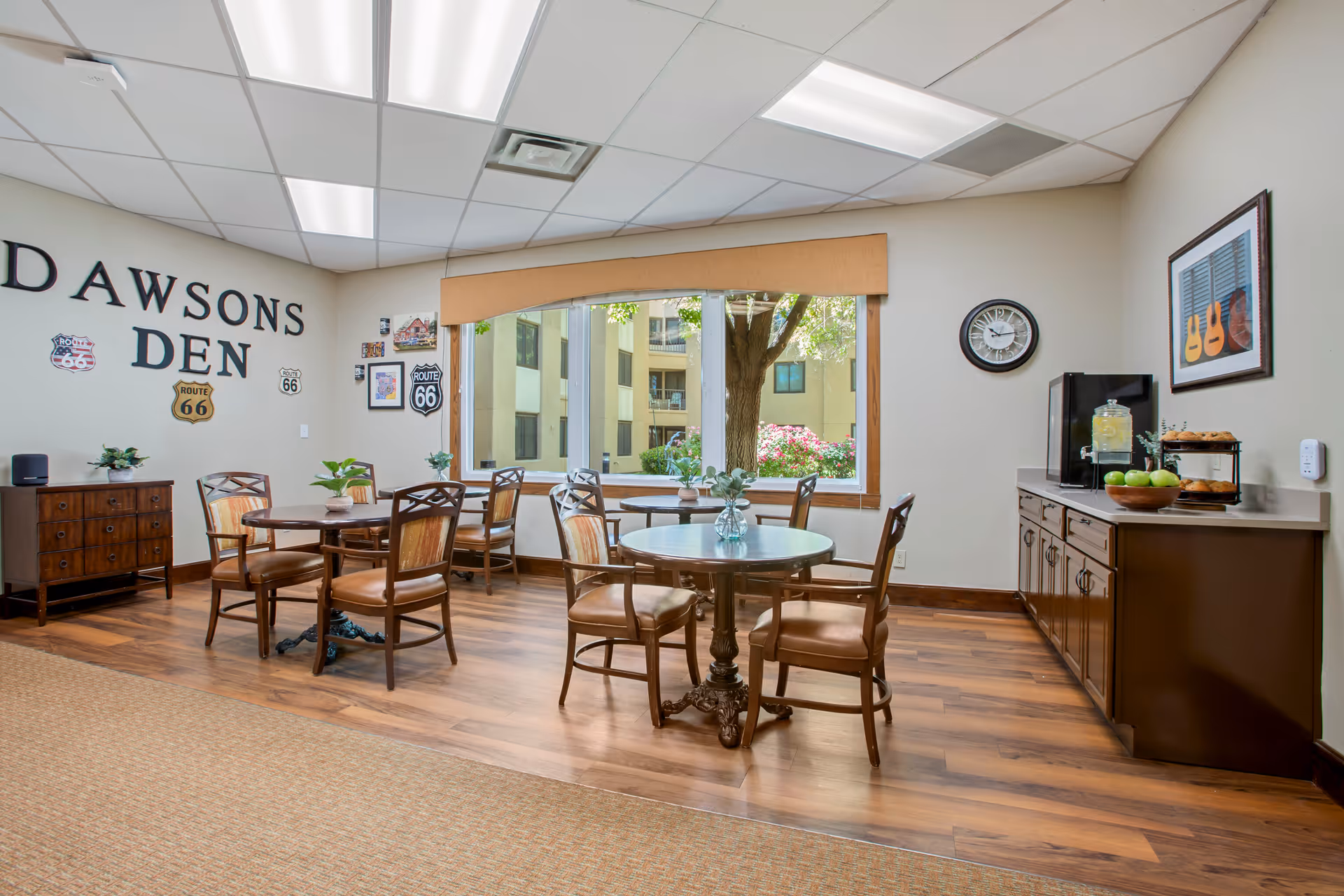A cozy common room named Dawsons Den with wooden floors, round tables and chairs, a large window showing an outdoor view with trees and flowers, wall decorations including Route 66 signs, a clock, and a sideboard with a water dispenser, fruit bowl, and baked goods.