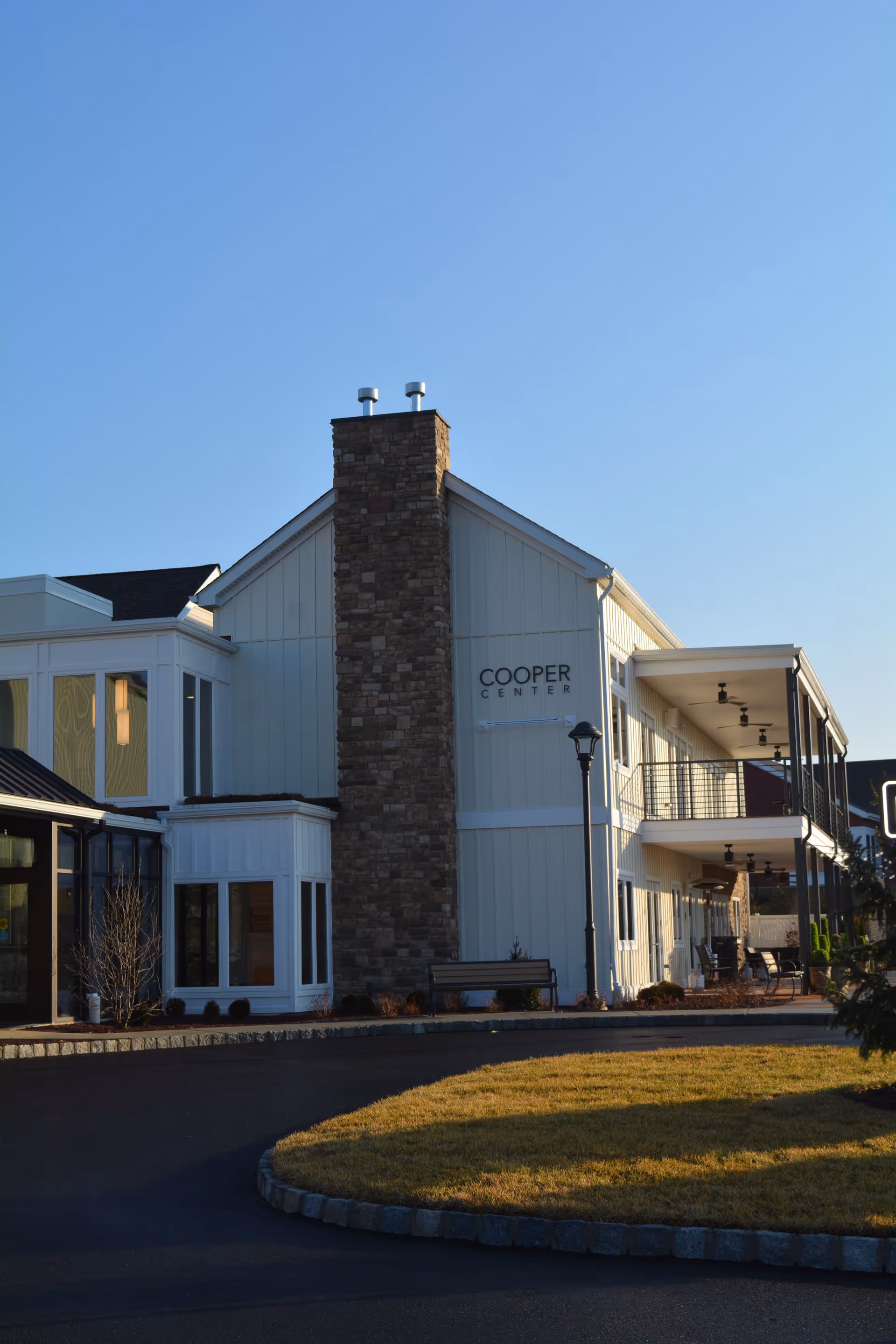 Exterior view of a two-story building named Cooper Center with white siding and a stone chimney, surrounded by a paved driveway and a small grassy area under a clear blue sky.