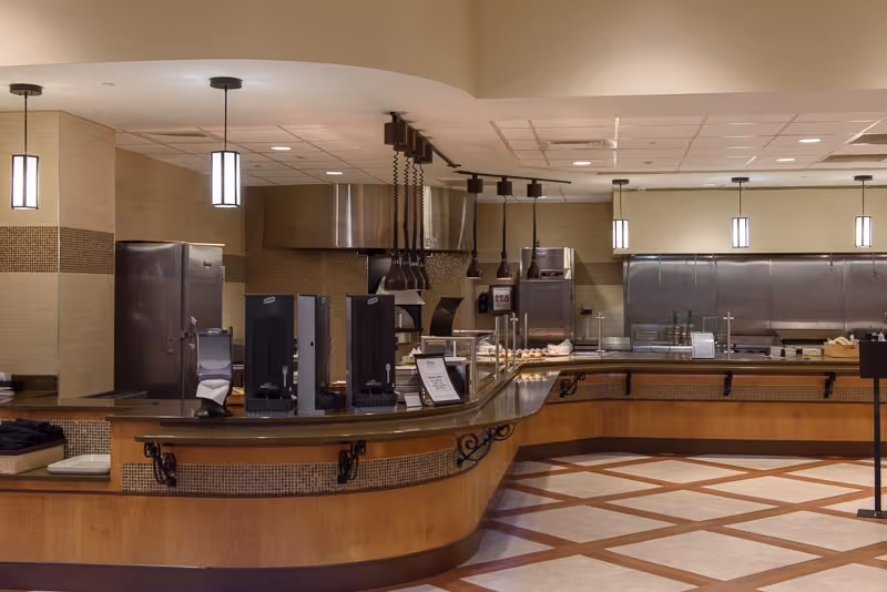 Empty cafeteria-style serving counter with hanging pendant lights and a stainless-steel commercial kitchen in the background.