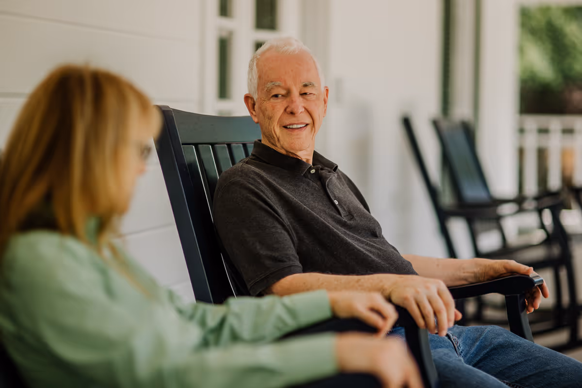 An elderly man and a woman sitting and talking on black rocking chairs on a porch with white walls and windows in the background.