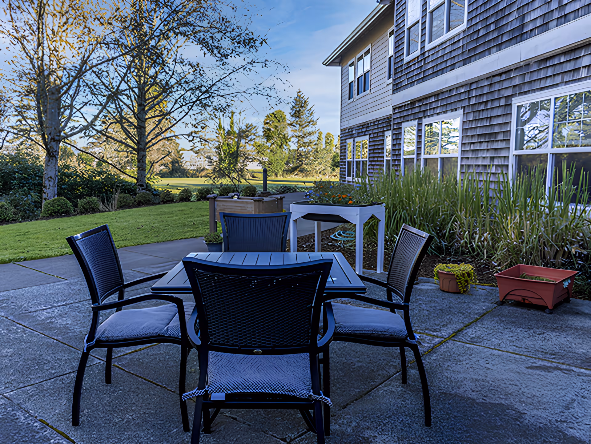 Outdoor patio area with a square table and four chairs on a concrete surface next to a building with multiple windows. There are plants and trees surrounding the patio and a grassy lawn extending into the background under a clear blue sky.