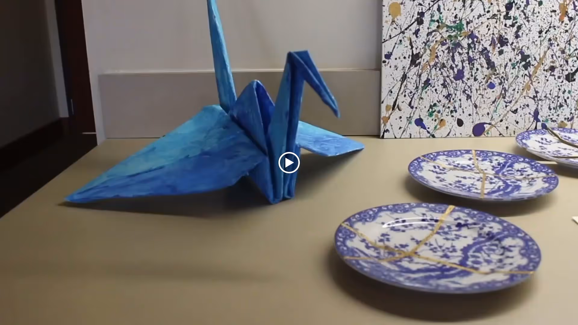 A blue origami crane displayed on a beige surface next to three decorative plates with blue floral patterns and gold repair lines, with an abstract splatter painting in the background.