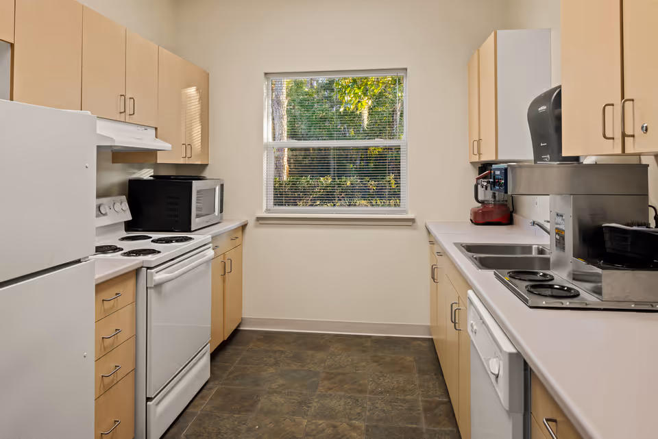 A clean and well-lit kitchen with light wood cabinets, a white refrigerator, electric stove, microwave, dishwasher, and a double sink. A window with blinds shows greenery outside. The countertops are light-colored, and the floor has dark tiles.