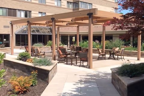 Outdoor patio area at a senior living facility with a wooden pergola, several chairs and tables arranged underneath, surrounded by raised garden beds with plants and shrubs, and a multi-story building in the background.