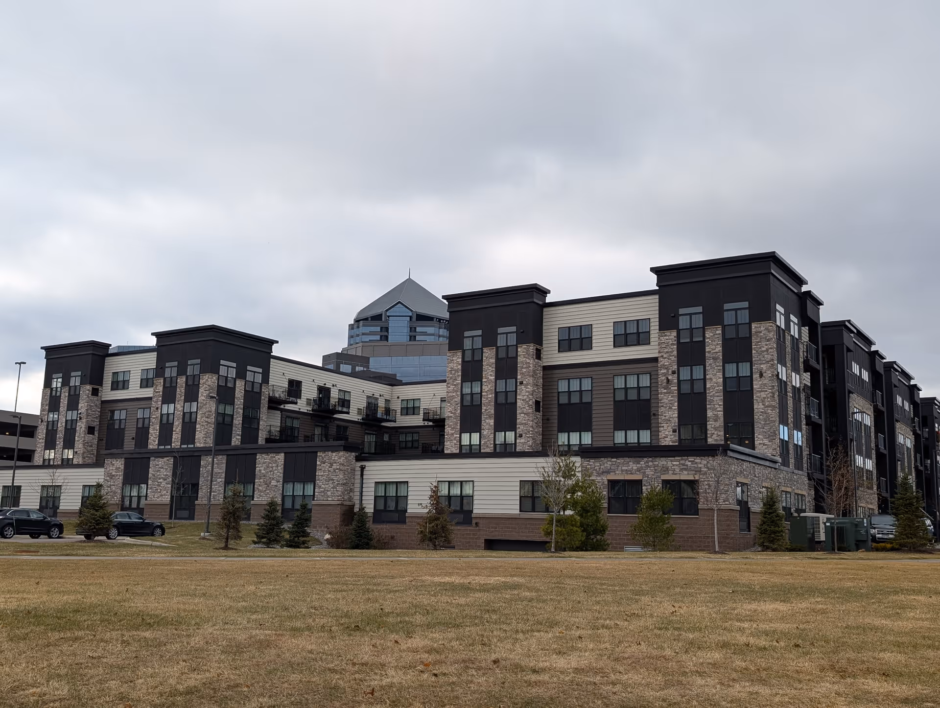Exterior view of a modern multi-story building with a combination of stone and dark paneling on the facade, surrounded by a grassy area and small trees under a cloudy sky.