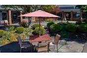 Outdoor patio area at Pinecrest Manor with a square wooden table, four chairs, and a large pink umbrella. The patio is surrounded by neatly trimmed bushes and greenery, with the building visible in the background.
