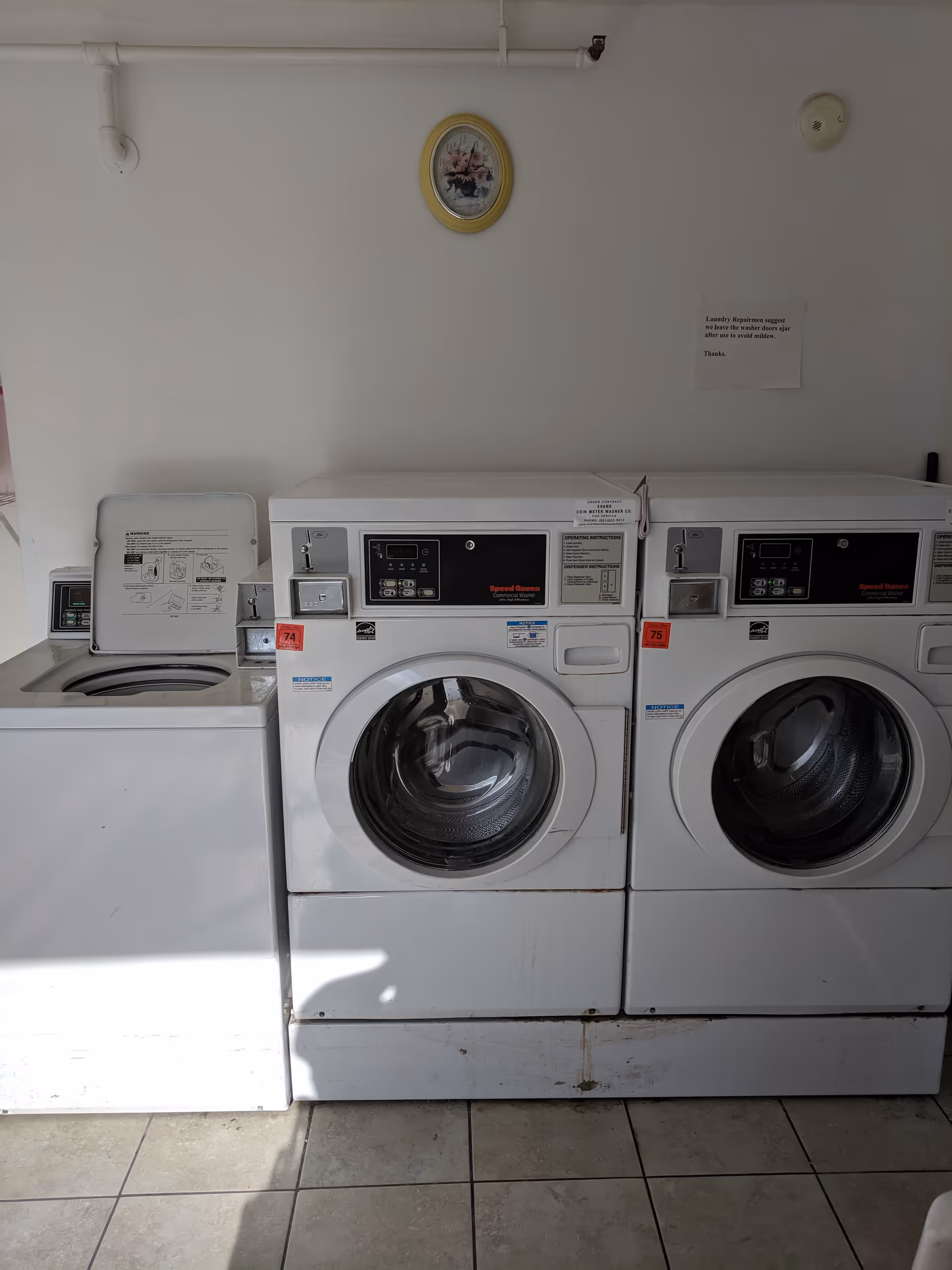Laundry room with three white commercial washing machines against a white wall. Above the machines, there is a small framed picture and a sign with laundry instructions. The floor is tiled.